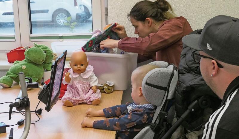 Ein Kind kann ein Feld auf dem Desktop mit den Augen fixieren – und der Computer spricht das gewünschte Wort aus. 
Der Kleine hat «Puppe anziehen» gewählt. Die Mutter sucht passende Kleider.